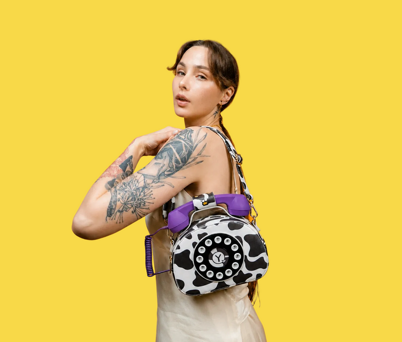 A woman with arm tattoos models a small vintage rotary phone-shaped shoulder bag with a black and white cow print pattern and purple accents, set against a solid orange background.